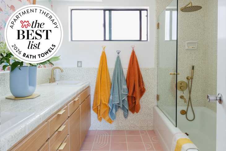 Bright bathroom featuring a light wood vanity, colorful towels hanging, and a glass shower with gold fixtures. Seal in upper left: "Apartment Therapy The Best List Bath Towels 2026"