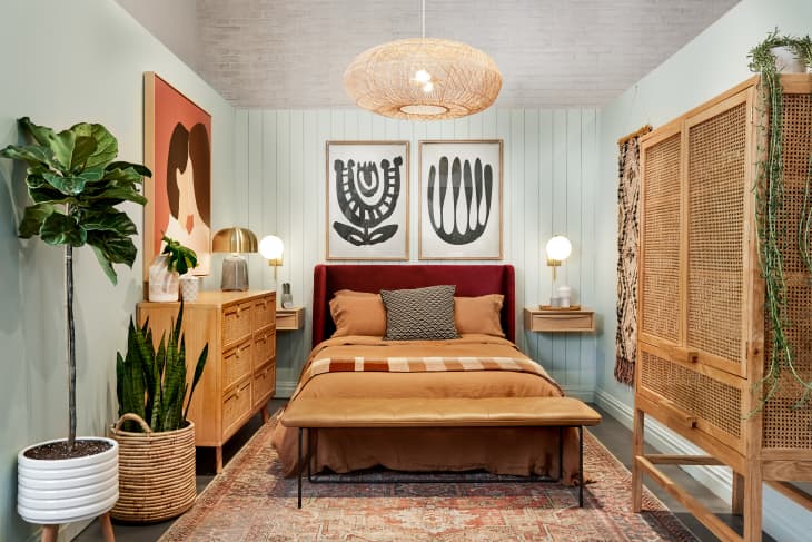 room with pale blue walls, graphic artwork, a rattan dresser, warm colored bed linens, snake plant on ground in basket