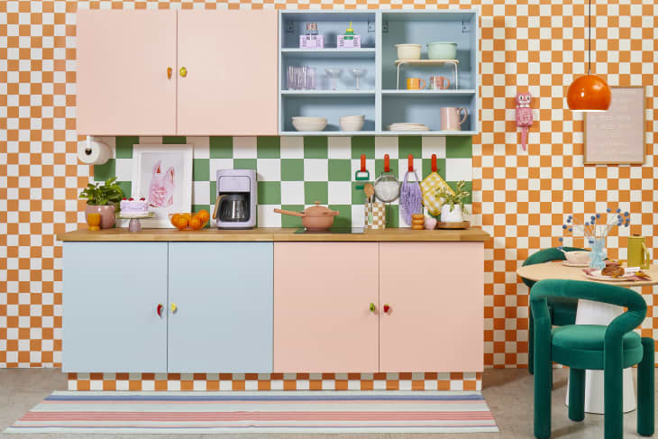 Head on view of a small kitchen with colorful cabinetry, orange and white checkered wallpaper, and a green and  white checkered backsplash.
