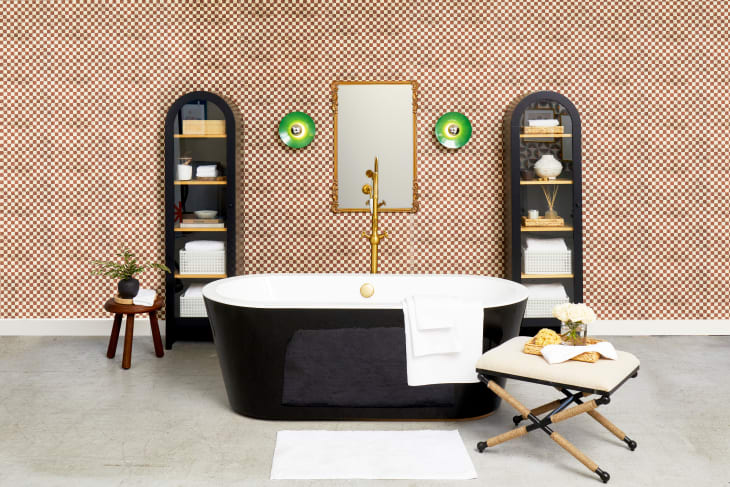Head on view of a black bathtub, with gold finishes in a bathroom with beige and brown checkered wallpaper, green circular sconces and a rectangular gold framed mirror, and a beige stool to the right of the bathtub. On either side of the mirror are black armoires with glass doors.