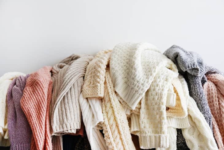 A bunch of light-colored sweaters draped over the top of a piece of furniture. White wall in background.