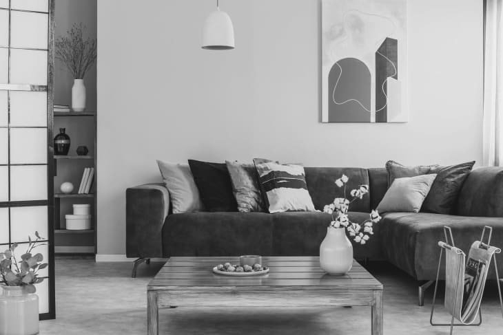 Grayscale photo of a square, wooden table standing in front of a brown, suede sofa with decorative cushions in cozy living room interior