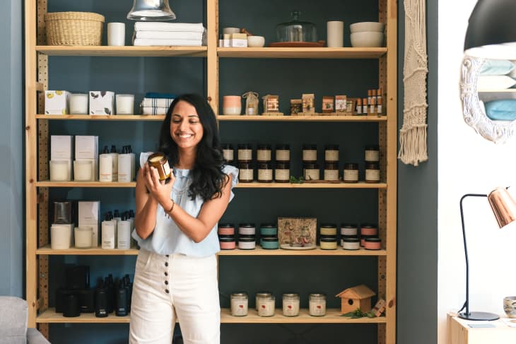 woman reading label on candle in store. There are shelves behind her with many more candles and other small household goods