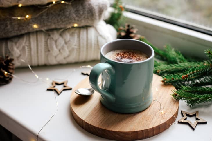 Cozy home picture of blue ceramic cup with coffee on window sill, Christmas decorations, warm knitted sweaters and pine tree green branches in background