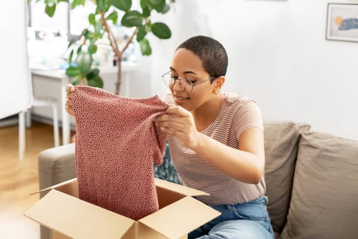 woman opening a cardboard box and pulling out a shirt. she is sitting on a sofa. tree/plant in background