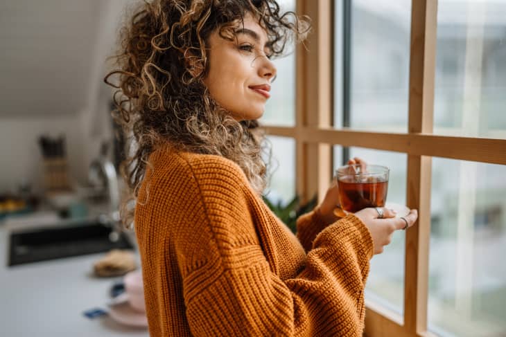woman stands in front of a window looking out with slight smile. She is wearing a cozy oversized burnt orange sweater and holding a glass mug of tea. Kitchen in background