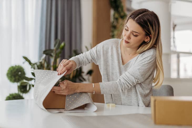 Woman at white table wrapping a gift with black and cream fabric or paper. plants and window in background