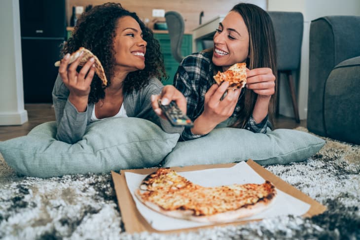 Two young women eating pizza and watching TV together at home.