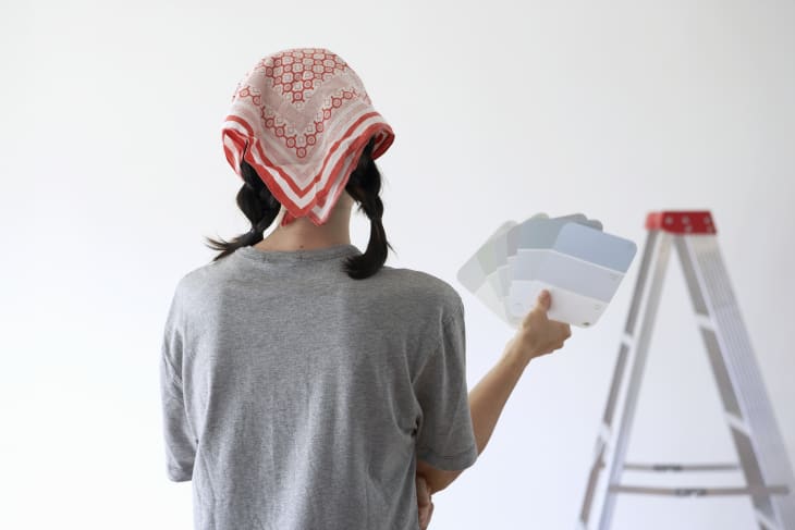 Woman holding assorted color swatches standing in front of a wall with a ladder, rear view