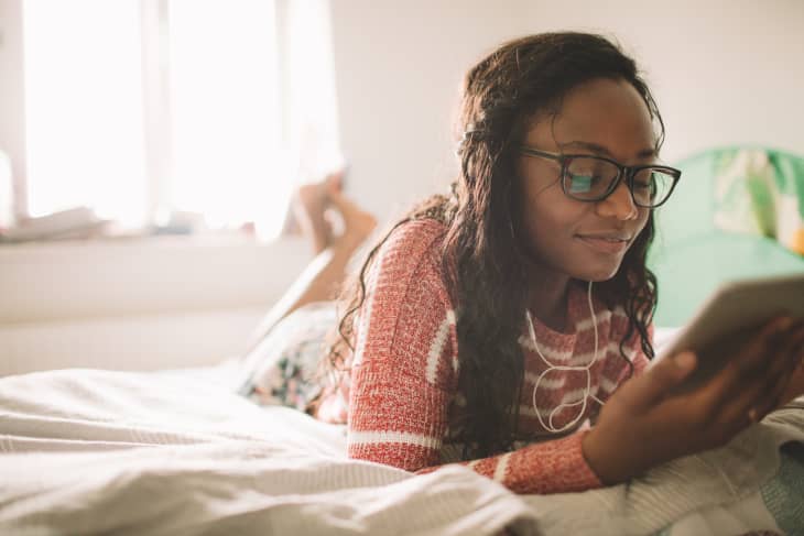 Photo of a young woman using her digital tablet while resting on the bed in her bedroom