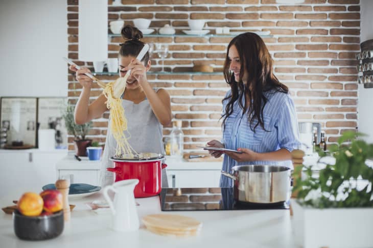 2 women in a kitchen. one is cooking spaghetti and lifting it out of a red stock pot. the other one is watching while holding a tablet. White countertops and brick wall in background