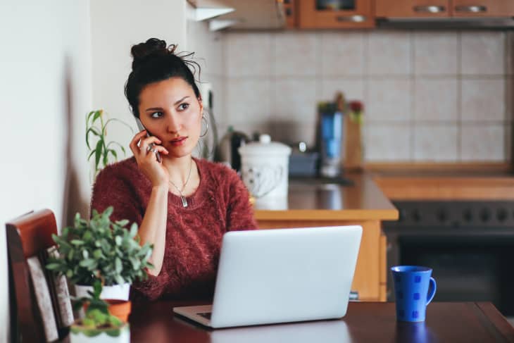 woman sitting in kitchen with a laptop. She is on the phone and has a blue coffee mug
