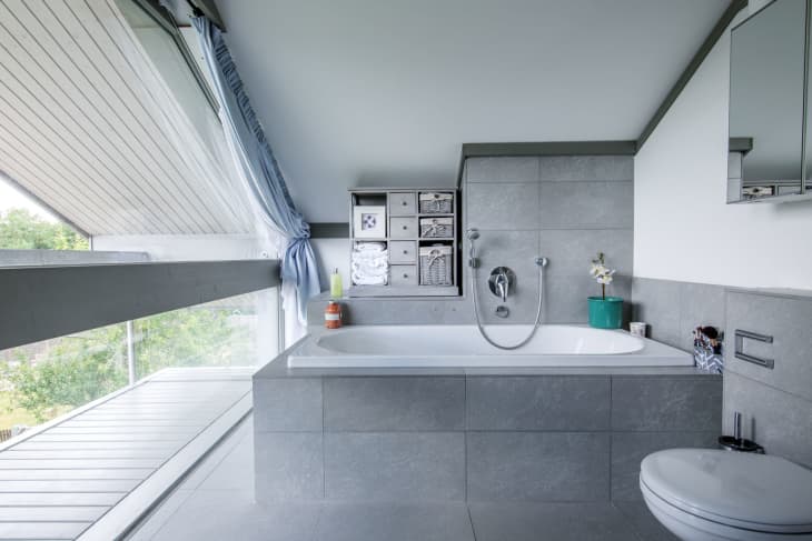 Modern bathroom with gray tiles, a bathtub, wall shelves with towels, a potted plant, and a large window with blue curtains.