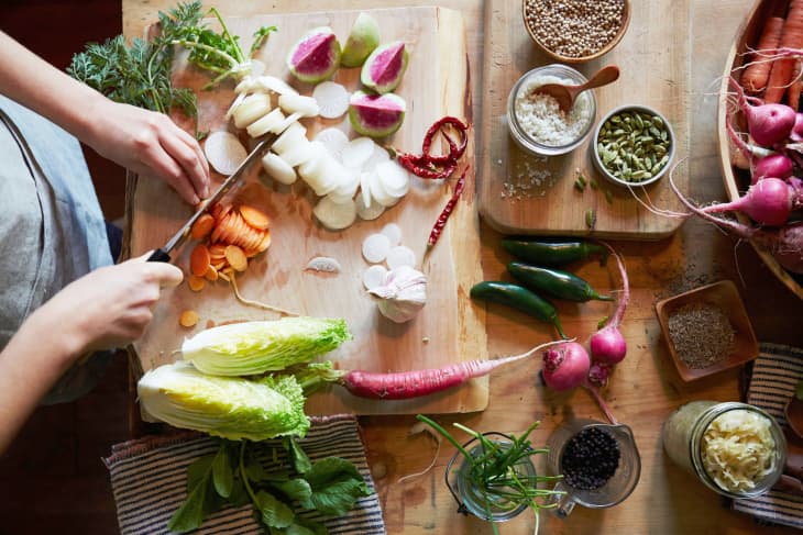 Person slicing vegetables on a wooden board with radishes, carrots, spices, and herbs on a kitchen table.