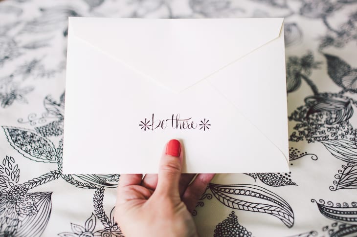 Hand holding an invitation envelope with hand lettering that reads "be there"