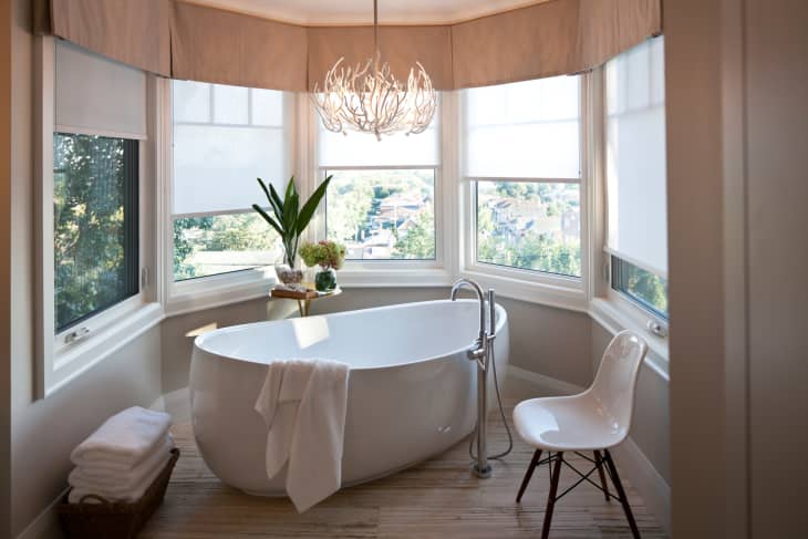 Freestanding bathtub in a bay window bathroom with a modern chair, plant, and chandelier.