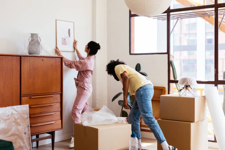 Young woman hanging framed picture on wall near boyfriend during relocation into new apartment