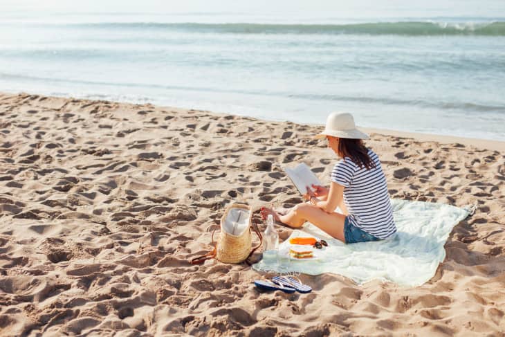Woman in striped shirt and hat reading on a beach blanket with a straw bag, flip-flops, and snacks nearby.