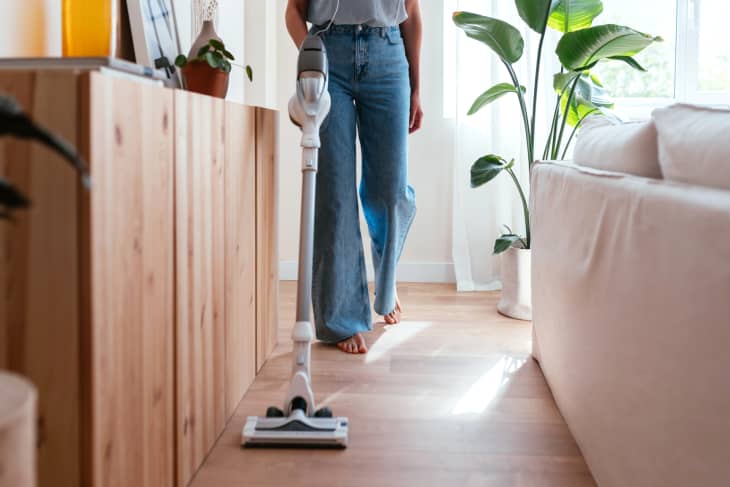 person in jeans vacuuming laminate floor, window and tall plant behind her