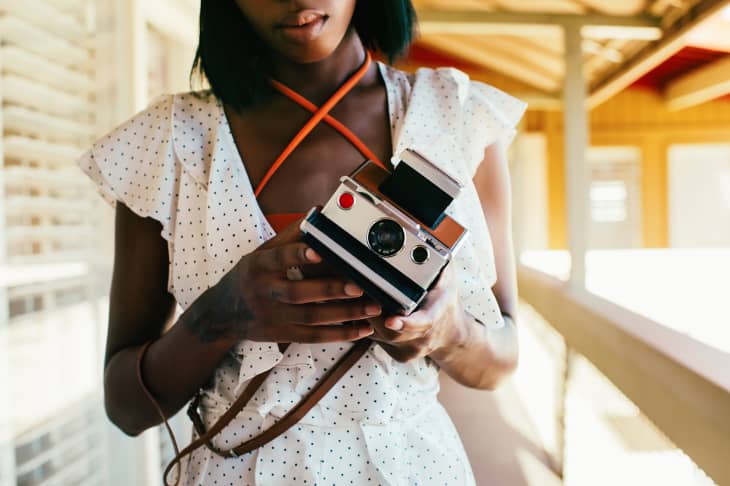 woman wearing white dress with small black polka dots holding white polaroid camera on a brown leather strap