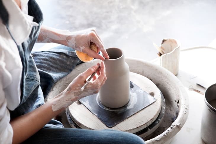 Person painting a clay pot they've made on a pottery wheel. Their hands are covered with drying clay.
