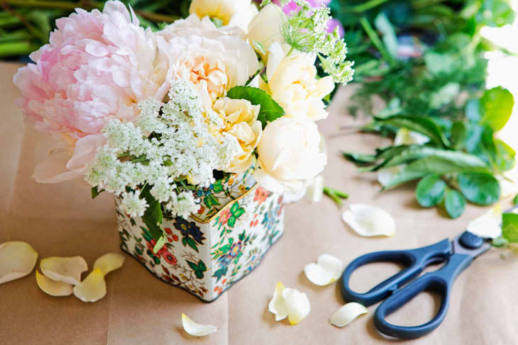 Floral arrangement with pink peonies, white roses, and greenery in a patterned vase, surrounded by petals and scissors.