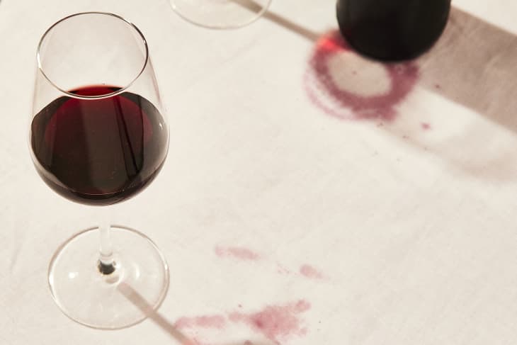 Glass of red wine on off-white tablecloth. There are wine stains on the tablecloth fabric, and a wine bottle can be seen in the upper right corner