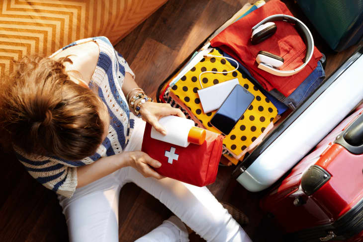 Upper view of modern woman in white pants and striped blouse at modern home in sunny summer day packing first aid kit and SPF in open travel suitcase.