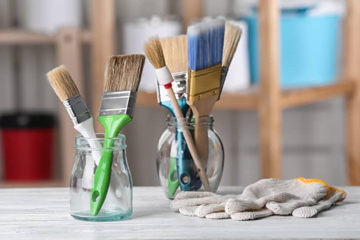 a variety of used paint brushes in glass jars. a pair of gloves sits on the table next to them