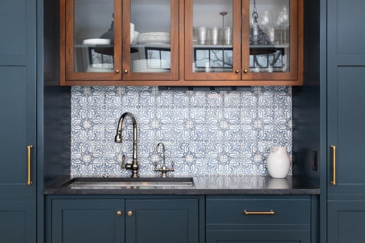 A kitchen sink with a beautiful pattern tiled backsplash with a chrome faucet, black granite countertops, and surrounded by blue and wood cabinets