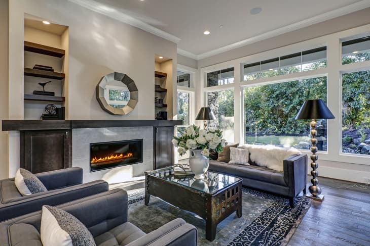 living room filled with built-in cabinets flanking round mirror atop grey tile fireplace, tufted sofa facing two armchairs and window wall overlooking lush outdoors