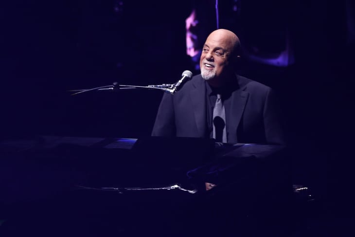 Billy Joel at piano in all black suit with blue and purple lighting
