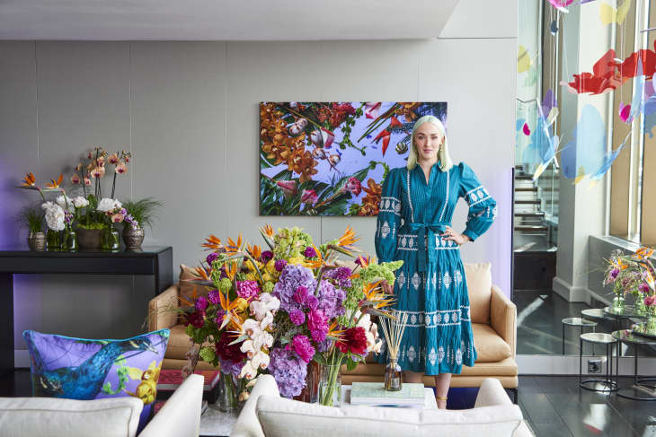 Woman in a blue dress stands in a colorful living room with vibrant floral arrangements and abstract art.