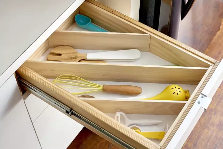 Open kitchen drawer with wooden dividers holding utensils, including a yellow whisk, wooden spatula, and citrus juicer.