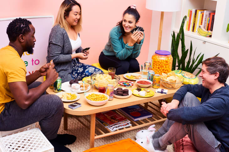 Four friends playing cards around a coffee table with snacks, drinks, and a jar of cheese balls in a cozy living room.