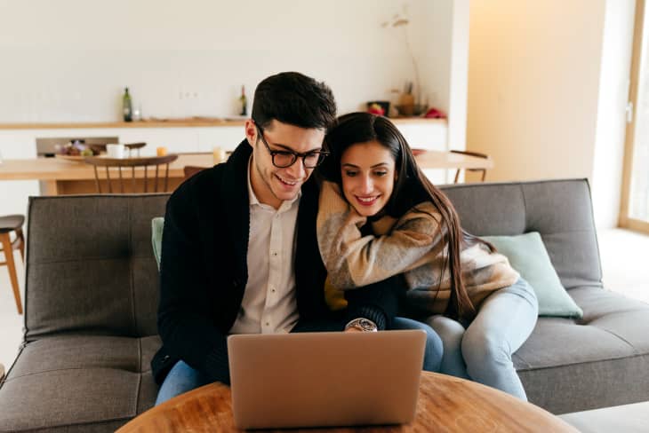 Couple sitting on a gray sofa using a laptop in a cozy living room with a wooden dining table in the background.