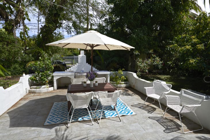 Outdoor patio with white wicker chairs, wooden table, large umbrella, and a blue patterned rug surrounded by lush greenery.