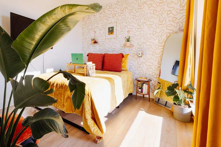Cozy bedroom featuring a yellow bedspread, red pillows, a green laptop, and large plants near a mirror and window.