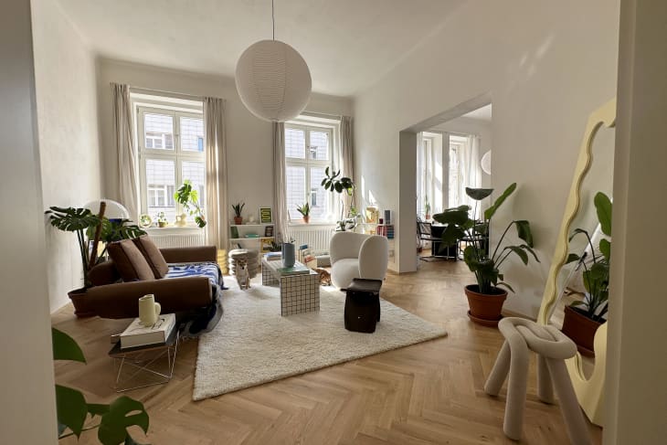 Living room with cream shag rug, white tile coffee table with colorful hardback coffee table books, wood herringbone floors, white walls, large windows with white curtains and plants around, checkered dog bed in front of window, white shelves with books, modern/80s looking art objects, brown loveseat with blue and white throw, white round armchair, full length wavy mirror with gold frame,