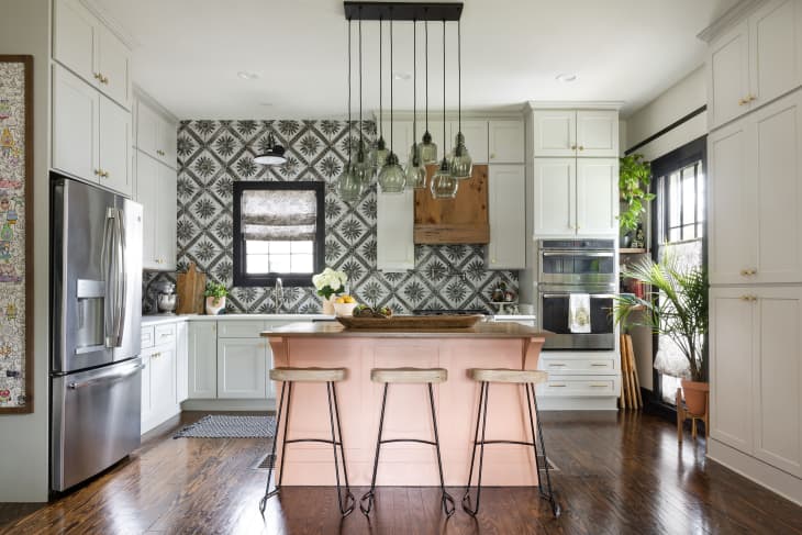 Kitchen with pale peach colored island with 3 stools, white walls with one black and white patterned wallpaper wall, pendant lamp with multiple lights, wood floor, white cabinets