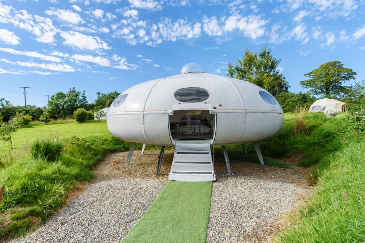 Futuristic UFO-shaped cabin with open door, set in a grassy field under a blue sky.