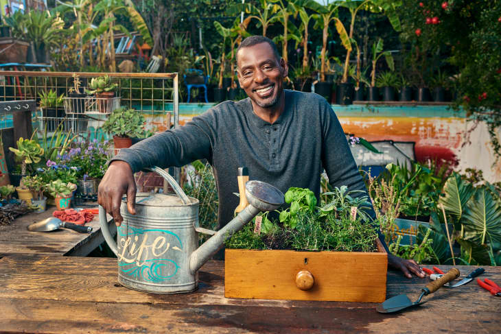Man smiling at a garden table with a watering can and a wooden planter box filled with herbs and plants.
