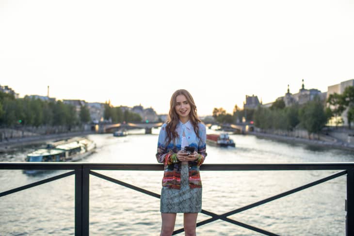 Woman in colorful outfit standing on a bridge over a river at sunset, with boats and cityscape in the background.