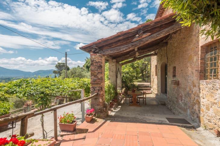Rustic stone patio with wooden pergola, surrounded by potted flowers and lush greenery, overlooking distant hills.