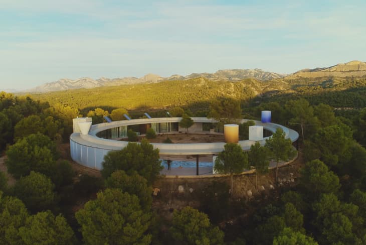 Circular modern building surrounded by dense forest with mountains in the background.