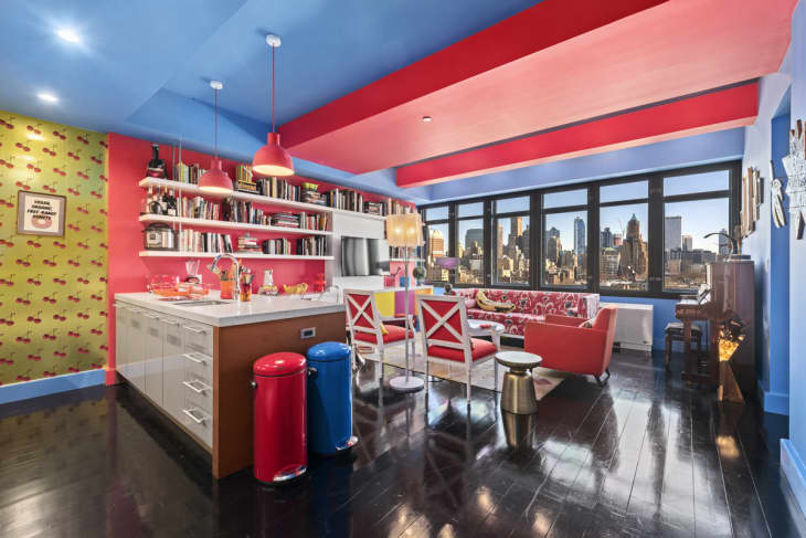 Colorful kitchen and living area with red and blue accents, cherry wallpaper, bookshelves, and city skyline view.