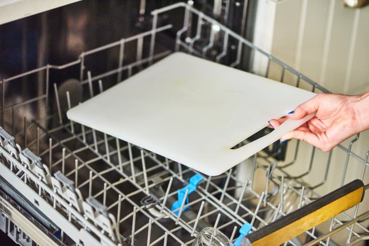 Hand placing a white cutting board into an open dishwasher rack.
