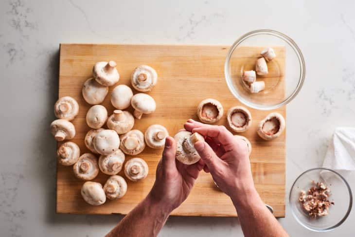 Hands preparing mushrooms on a wooden cutting board, with stems removed and placed in a glass bowl.