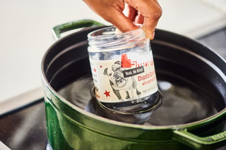 Hand placing a jar labeled "Chipotle Caesar" into a green pot filled with water on a stove.
