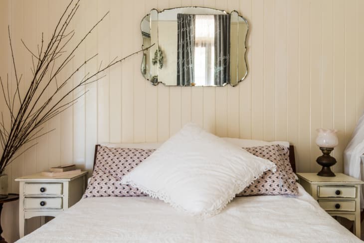 Bedroom with a white bed, patterned pillows, vintage mirror, and bedside tables with lamps and decor.